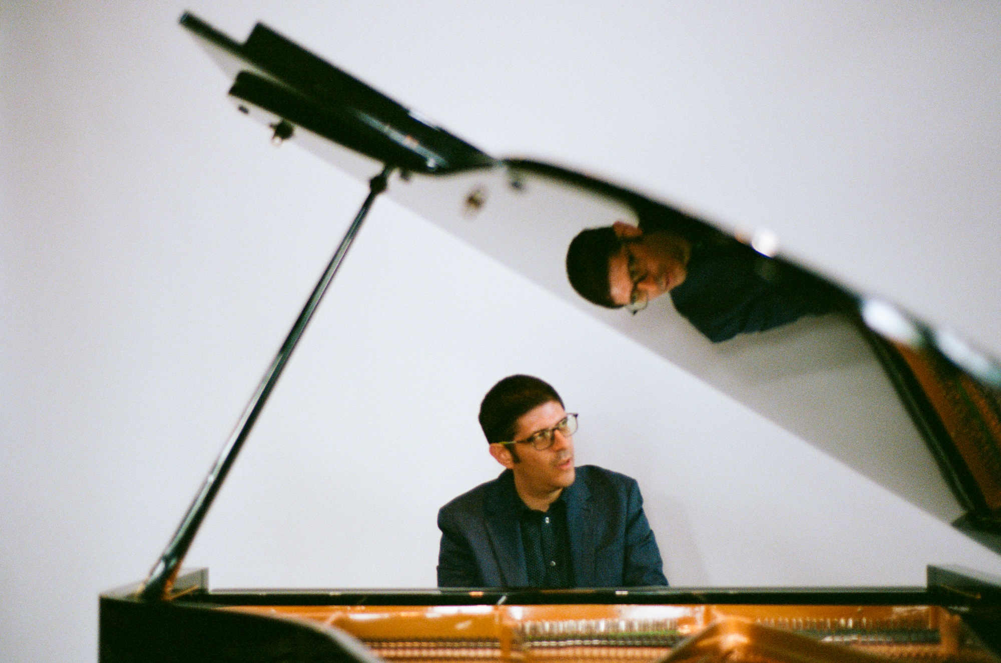 Robin Green at a grand piano with the lid open, looking slightly left. A white wall backdrop sits behind him.